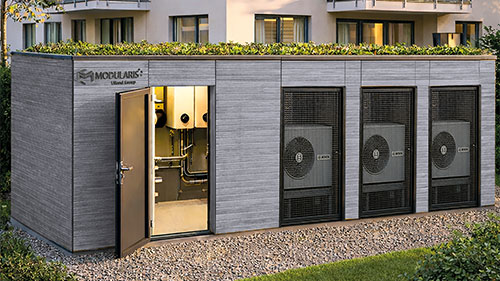Modularis Energiezentrale mit grauer Fassade, Gründach und drei integrierten Wärmepumpen vor einem Mehrfamilienhaus