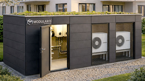 Modularis Energiezentrale mit dunkler Fassade, Gründach und zwei integrierten Wärmepumpen vor einem Mehrfamilienhaus